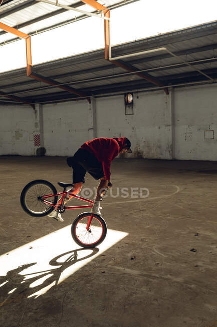 Vista laterale di un giovane caucasico in equilibrio sulla ruota anteriore di una bici BMX in un pozzo di luce solare mentre pratica trucchi in un magazzino abbandonato — Foto stock