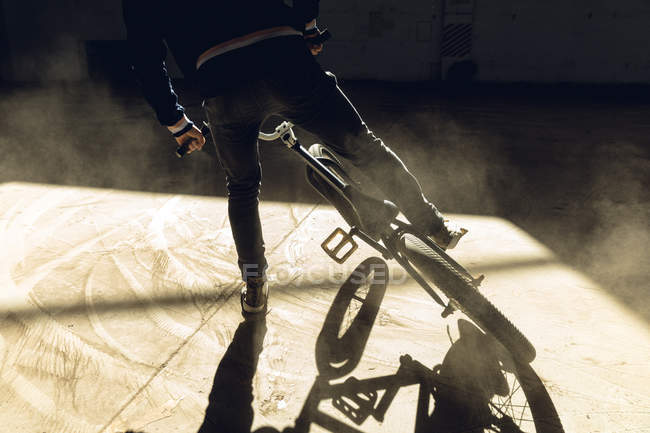Вид сзади низкая часть человека скользит боком к остановке на велосипеде BMX в валу солнечного света, практикуя трюки в заброшенном складе — стоковое фото