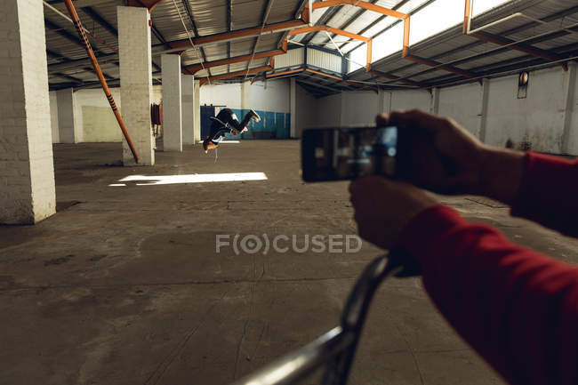 Vista lateral de un joven caucásico haciendo un salto mortal en un almacén abandonado, siendo filmado en un teléfono inteligente por su amigo sentado en una bicicleta BMX en primer plano - foto de stock
