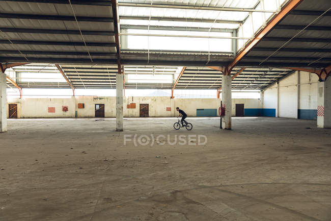 Side view of a young Caucasian man riding a BMX bike while practicing tricks in an abandoned warehouse — Stock Photo