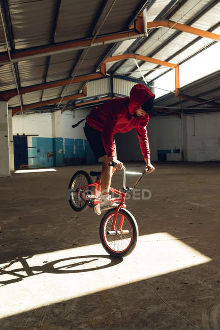 Nahaufnahme eines jungen kaukasischen Mannes, der auf dem Vorderrad eines BMX-Fahrrads in einem Sonnenlicht balanciert, während er in einer verlassenen Lagerhalle Tricks übt — Stockfoto