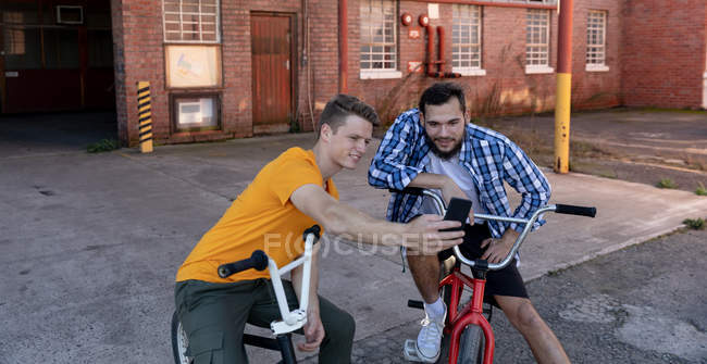 Nahaufnahme von zwei jungen kaukasischen Männern, die auf BMX-Fahrrädern sitzen und reden, wobei der eine dem anderen sein Smartphone vor einer verlassenen Lagerhalle zeigt. — Stockfoto