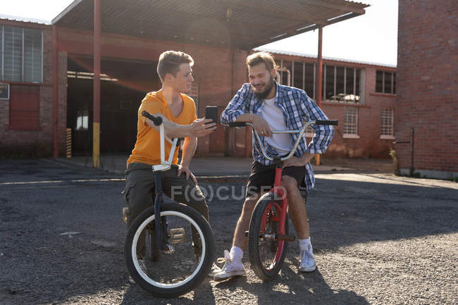 Nahaufnahme von zwei jungen kaukasischen Männern, die auf BMX-Fahrrädern sitzen und reden. Einer zeigt dem anderen sein Smartphone vor einer verlassenen Lagerhalle, die von Sonnenlicht beleuchtet wird. — Stockfoto