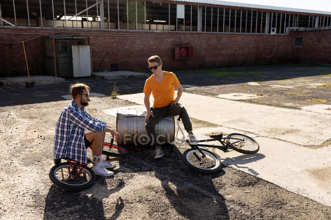 Seiten- und Frontansicht von zwei jungen kaukasischen Männern mit Sonnenbrille, die mit BMX-Fahrrädern vor einer verlassenen Lagerhalle in der Sonne sitzen und reden — Stockfoto