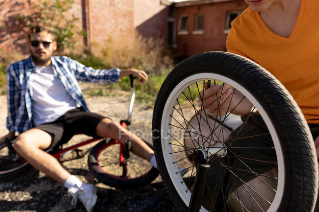 Vista frontal de cerca de dos jóvenes caucásicos sentados con bicicletas BMX fuera de un almacén abandonado al sol, uno en primer plano revisando un neumático y otro en el fondo usando gafas de sol y sentado en su bicicleta - foto de stock