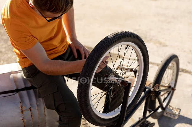 Vista lateral del hombre con gafas de sol sentado haciendo mantenimiento a la rueda de su bicicleta BMX fuera en el sol - foto de stock