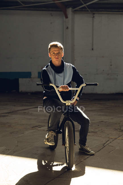 Retrato de perto de um jovem caucasiano sentado em uma bicicleta BMX olhando para a câmera em um armazém abandonado, iluminado pela luz solar — Fotografia de Stock