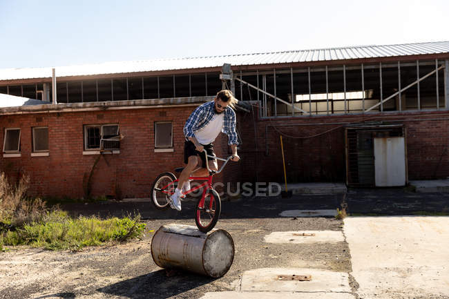 Side view of a young Caucasian man wearing sunglasses balancing on a barrel on the front wheel of a BMX bike outside an abandoned warehouse in the sun — Stock Photo
