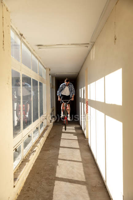 Вид на молодий Кавказький чоловік носіння сонцезахисні окуляри стрибки на BMX Bike у вузькому коридорі на занедбаному складі на сонці — стокове фото