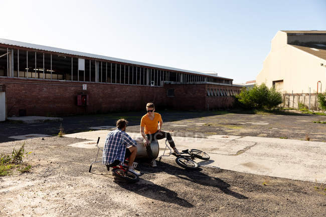 Rück- und Vorderansicht von zwei jungen kaukasischen Männern mit Sonnenbrille, die mit BMX-Fahrrädern vor einer verlassenen Lagerhalle in der Sonne sitzen und reden — Stockfoto