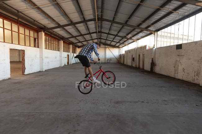 Vista lateral de un joven caucásico saltando en una bicicleta BMX en un almacén abandonado - foto de stock