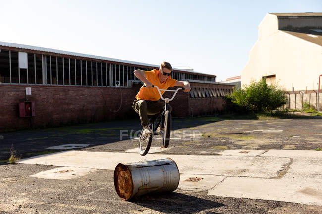 Front view of a young Caucasian man wearing sunglasses jumping over a barrel on a BMX bike outside an abandoned warehouse in the sun — Stock Photo