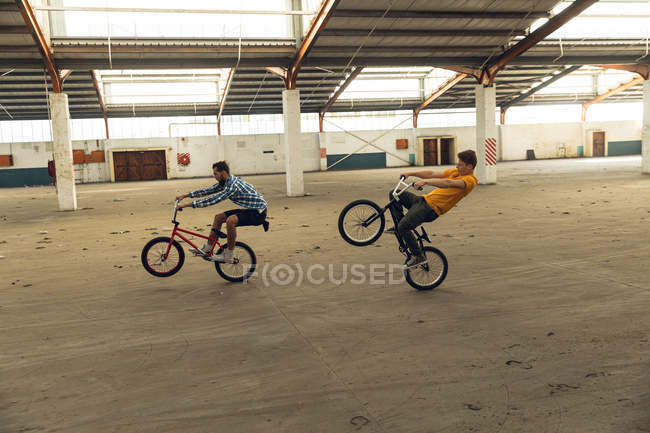Vista lateral de dos jóvenes caucásicos corriendo sobre las ruedas traseras de sus bicicletas BMX en un almacén abandonado - foto de stock
