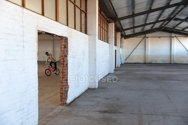 Side view of a young Caucasian man riding a BMX bike in an abandoned warehouse, seen through a broken doorway — Stock Photo