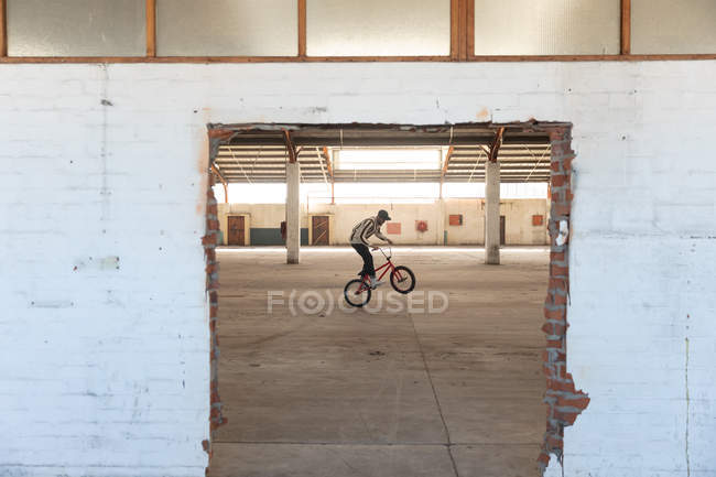 Vista lateral de um jovem caucasiano balanceando na roda traseira de uma bicicleta BMX enquanto pratica truques em um armazém abandonado, visto através de uma porta quebrada — Fotografia de Stock