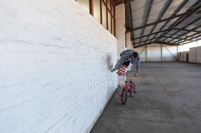 Vista frontal de un joven caucásico corriendo sobre una pared y sosteniendo los manillares de una bicicleta BMX en un almacén abandonado - foto de stock