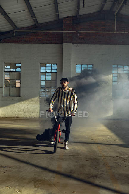 Frontansicht eines jungen kaukasischen Mannes auf einem BMX-Fahrrad mit einer grauen Rauchgranate daran in einer verlassenen Lagerhalle — Stockfoto