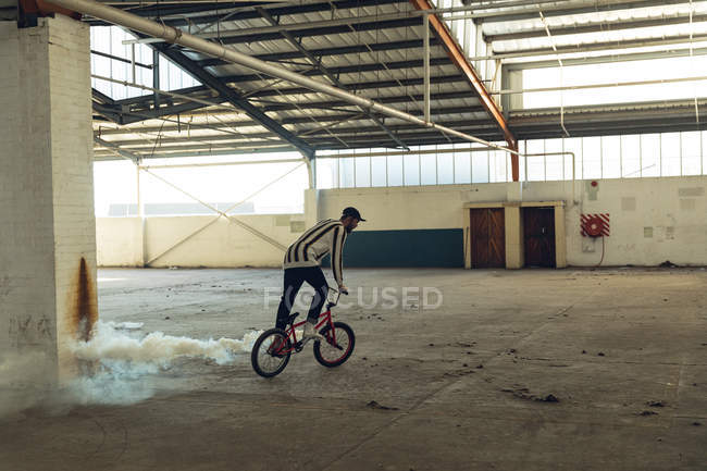Vista lateral de un joven caucásico con una gorra de béisbol montada en una bicicleta BMX con una granada de humo blanca en un almacén abandonado - foto de stock
