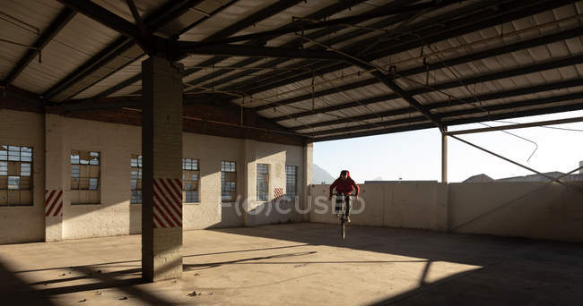 Vista frontal de um jovem caucasiano andando de bicicleta BMX e pulando enquanto pratica truques em um armazém abandonado — Fotografia de Stock