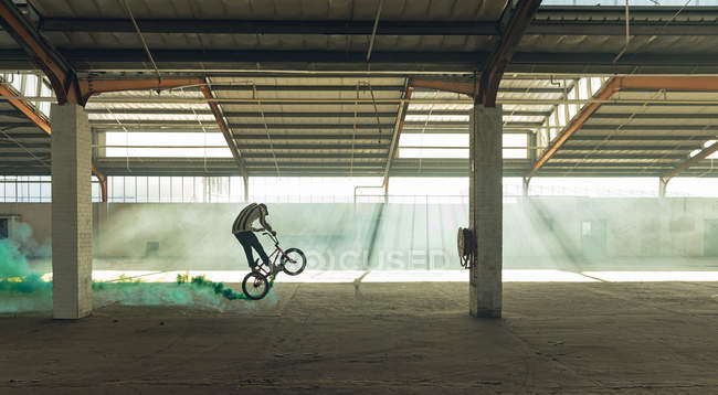 Вид сбоку на молодого кавказца в бейсболке и прыгающего на велосипеде BMX с зеленой дымовой гранатой на заброшенном складе — стоковое фото