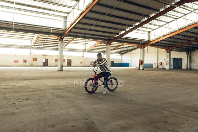 Portrait d'un jeune homme caucasien barbu assis sur un vélo BMX regardant la caméra dans un entrepôt abandonné — Photo de stock