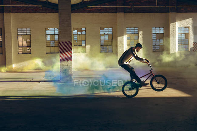 Seitenansicht eines jungen kaukasischen Mannes mit Baseballmütze, der auf dem Hinterrad eines BMX-Fahrrads reitet, an dem eine blaue Rauchgranate angebracht ist, in einer verlassenen Lagerhalle — Stockfoto