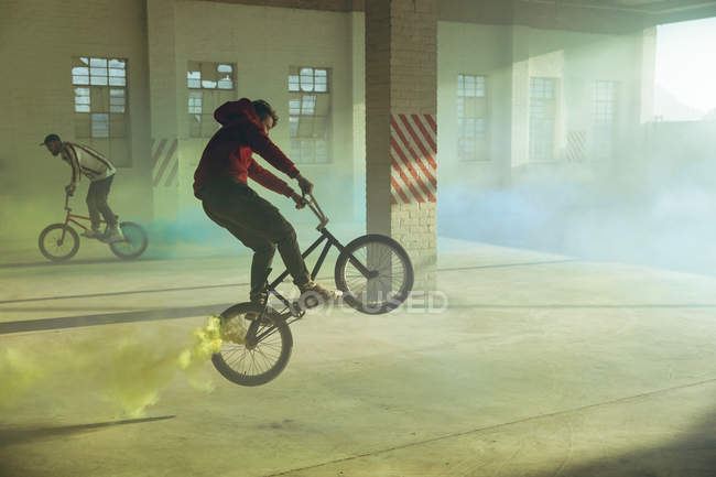 Vue latérale de deux jeunes hommes caucasiens chevauchant et faisant des tours sur des vélos BMX avec des grenades fumigènes jaunes et bleues attachées à eux, dans un entrepôt abandonné — Photo de stock