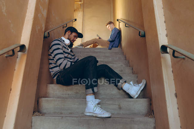 Seitenansicht von zwei hippen jungen kaukasischen Männern, die auf Stufen sitzen und Smartphones in einer verlassenen Lagerhalle benutzen — Stockfoto