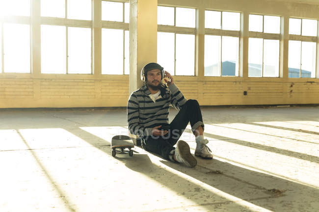 Front view of a hip young Caucasian man wearing a baseball cap and headphones sitting on the floor leaning against a pillar in an abandoned warehouse in the sun, holding a smartphone and listening to music, a skateboard beside him — Stock Photo