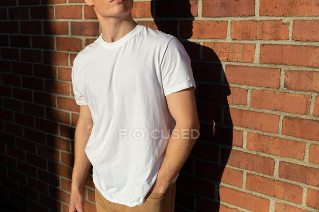 Vista lateral seção meio de um jovem caucasiano vestindo uma camisa branca em pé contra uma parede de tijolo ao sol olhando para longe — Fotografia de Stock