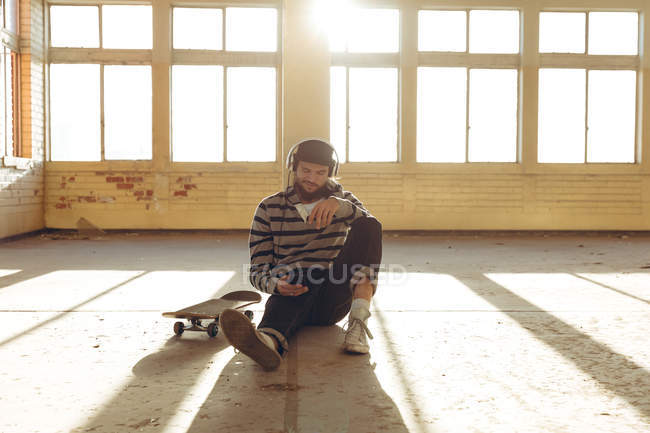 Front view of a hip young Caucasian man wearing a baseball cap and headphones sitting on the floor leaning against a pillar in an abandoned warehouse in the sun, holding a smartphone and listening to music, a skateboard beside him — Stock Photo