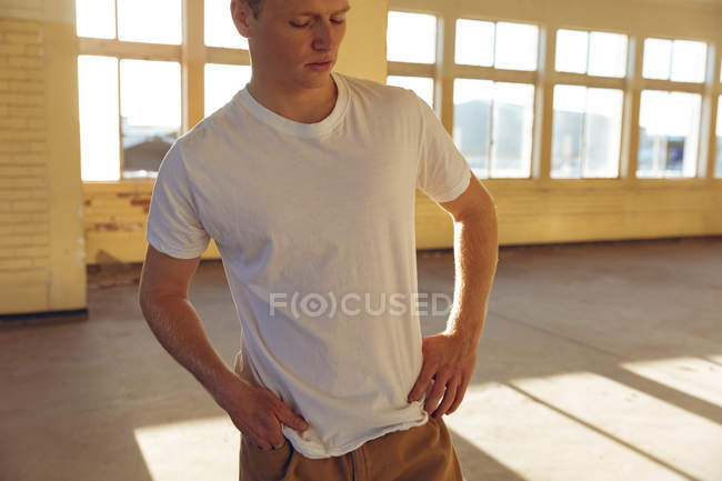 Vista frontale da vicino di un giovane caucasico che indossa una maglietta bianca in piedi in un magazzino abbandonato al sole, guardando in basso, con le mani sui fianchi — Foto stock