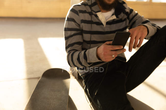 Vista frontal sección media de un joven caucásico sentado en el suelo en un almacén abandonado al sol, usando un teléfono inteligente, un monopatín a su lado - foto de stock