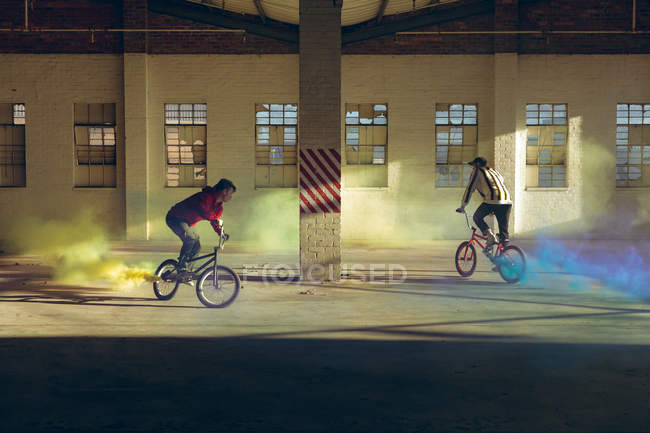 Seitenansicht von zwei jungen kaukasischen Männern, die mit gelben und blauen Rauchgranaten an ihren Fahrrädern durch Schächte des Sonnenlichts fahren, in einer verlassenen Lagerhalle — Stockfoto