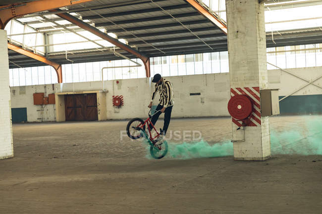 Side view of a young Caucasian man wearing a baseball cap riding and jumping on a BMX bike with a green smoke grenade attached to it, in an abandoned warehouse — Stock Photo