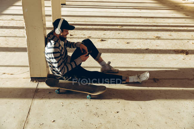 Side view of a hip young Caucasian man wearing a baseball cap and headphones sitting on the floor leaning against a pillar in an abandoned warehouse in the sun, holding a smartphone and listening to music, a skateboard beside him — Stock Photo