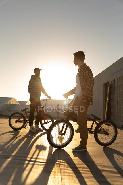 Vista lateral de cerca de dos jóvenes caucásicos de pie con bicicletas BMX y en la azotea de un almacén abandonado, retroiluminado por el sol poniente - foto de stock