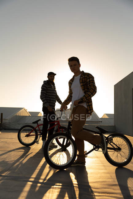 Вид сбоку на двух молодых кавказских мужчин, стоящих с велосипедами BMX и на крыше заброшенного склада, подсвеченного заходящим солнцем, смотрящих в камеру — стоковое фото