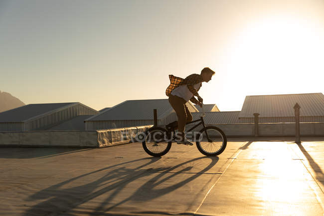 Vista lateral de un joven caucásico montando una bicicleta BMX en la azotea de un almacén abandonado, retroiluminado por el sol poniente - foto de stock