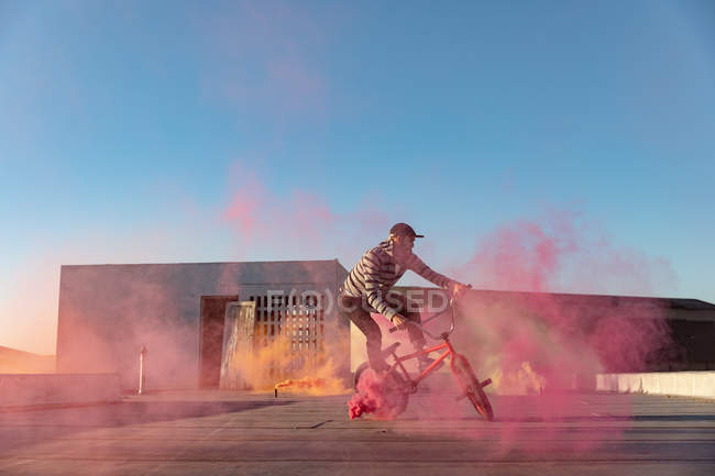 Side view of a young Caucasian man riding a BMX bike and doing tricks on the rooftop of an abandoned warehouse, with a pink smoke grenade attached to the bike — Stock Photo
