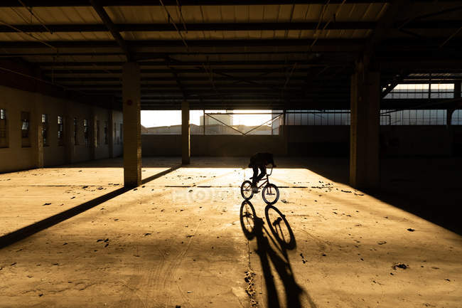 Vue latérale d'un jeune homme caucasien en vélo BMX et faisant des tours dans un entrepôt abandonné, rétroéclairé par la lumière du soleil — Photo de stock