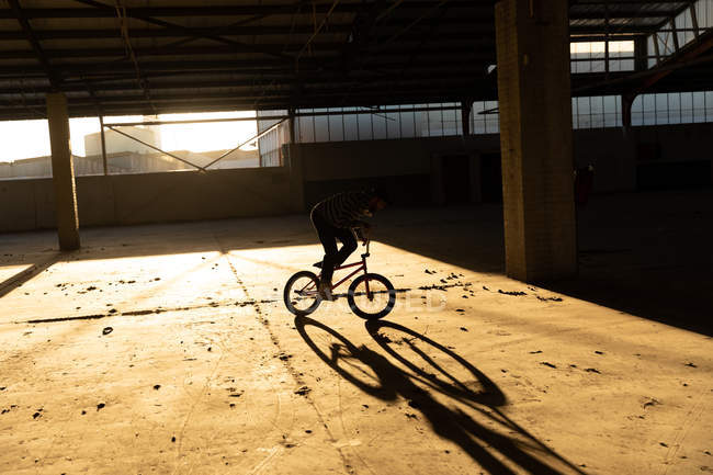 Vista lateral de um jovem caucasiano andando de bicicleta BMX enquanto pratica truques em um armazém abandonado, iluminado pela luz solar — Fotografia de Stock