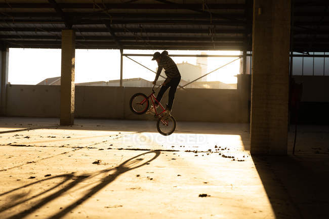Seitenansicht eines jungen kaukasischen Mannes, der auf einem BMX-Fahrrad springt, während er in einer verlassenen Lagerhalle im Gegenlicht der Sonne Tricks übt — Stockfoto