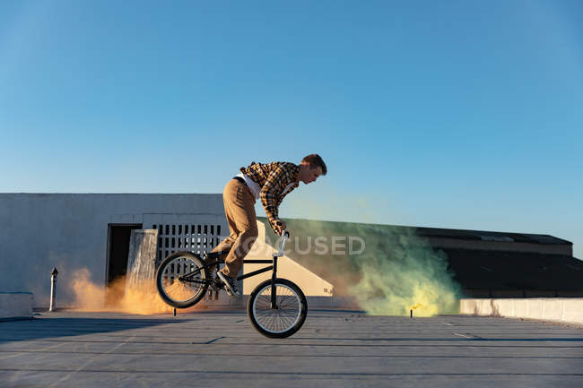Vue latérale d'un jeune homme caucasien se balançant sur la roue avant d'un vélo BMX faisant des tours sur le toit d'un entrepôt abandonné, avec des grenades fumigènes vertes et orange en arrière-plan — Photo de stock