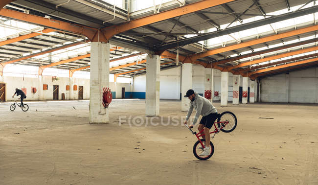 Side view of two young Caucasian men riding BMX bikes while practicing tricks in an abandoned warehouse, the rider in the foreground is balancing on the front wheel of his bike — Stock Photo