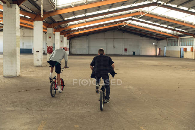 Vista trasera de dos jóvenes caucásicos montando bicicletas BMX mientras practican trucos en un almacén abandonado - foto de stock
