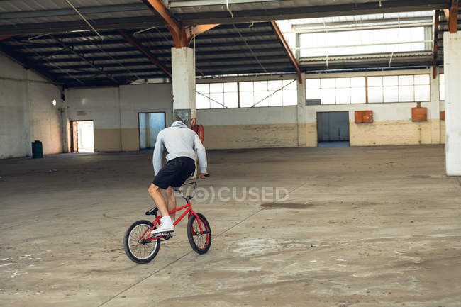 Vista trasera de un joven caucásico con pantalones cortos y una sudadera con capucha practicando trucos en una bicicleta BMX en un almacén abandonado - foto de stock