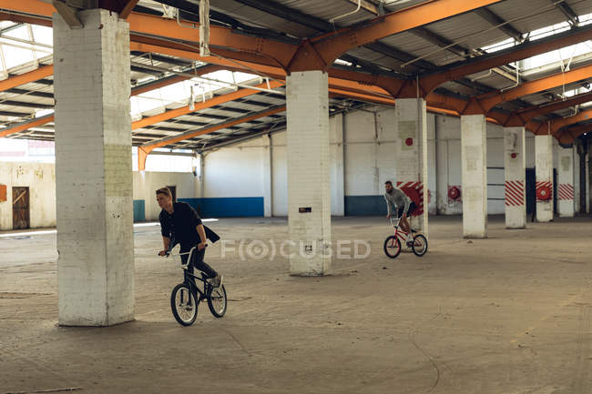 Vista frontal de dois jovens caucasianos andando de bicicleta BMX enquanto pratica truques em um armazém abandonado — Fotografia de Stock
