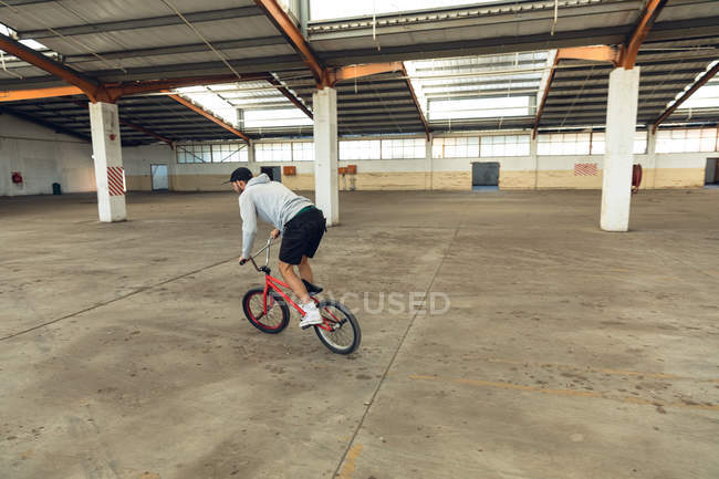 Vista lateral de um jovem caucasiano vestindo um boné de beisebol, shorts e um top com capuz andando de bicicleta BMX enquanto pratica truques em um armazém abandonado — Fotografia de Stock