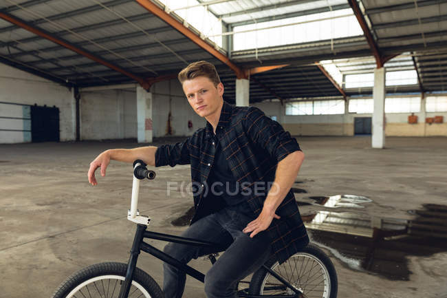 Вид збоку крупним планом молодого Кавказького чоловіка, одягнений в чорний, сидячи на велосипеді BMX і звертаючись дивитися на камеру в покинутому складі — стокове фото